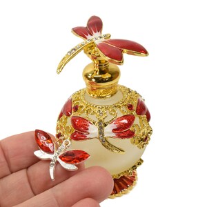 May include: A gold and white glass perfume bottle with a red and white dragonfly design. The bottle has a gold stopper with a red dragonfly on top. A smaller red and white dragonfly charm is held in a hand.