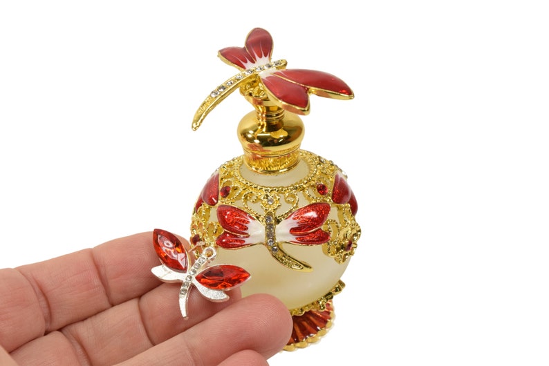 May include: A gold and white glass perfume bottle with a red and white dragonfly design. The bottle has a gold stopper with a red dragonfly on top. A smaller red and white dragonfly charm is held in a hand.