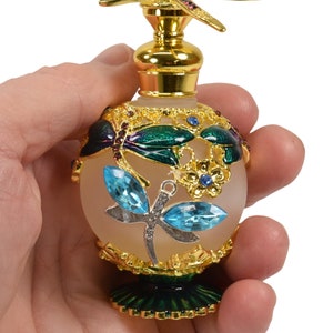 May include: A gold and green glass perfume bottle with a dragonfly design. The bottle has a frosted glass body and a gold metal cap with a dragonfly on top. The bottle is decorated with blue and green enamel.