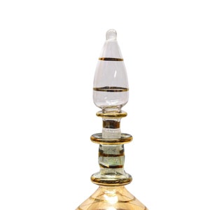 Puede incluir: Una botella de vidrio transparente con un tapón y una base dorados. La botella tiene un cuerpo redondo y un cuello largo y estrecho.