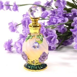 Puede incluir: Un pequeño frasco de perfume adornado con un tapón de vidrio transparente y una base de metal dorado. El frasco está decorado con flores moradas y hojas verdes. El frasco está sobre una superficie blanca.
