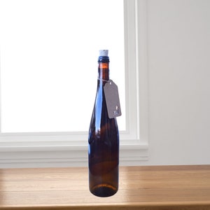 Puede incluir: Una botella de vidrio marr&oacute;n oscuro con un tap&oacute;n de corcho y una etiqueta de papel marr&oacute;n. La botella est&aacute; sobre una superficie de madera.