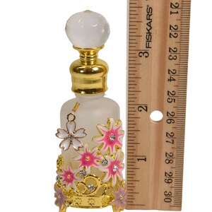 May include: A frosted glass bottle with a gold top and a clear crystal stopper. The bottle is decorated with pink and white flowers and rhinestones. The bottle is sitting on a gold metal stand with three legs.