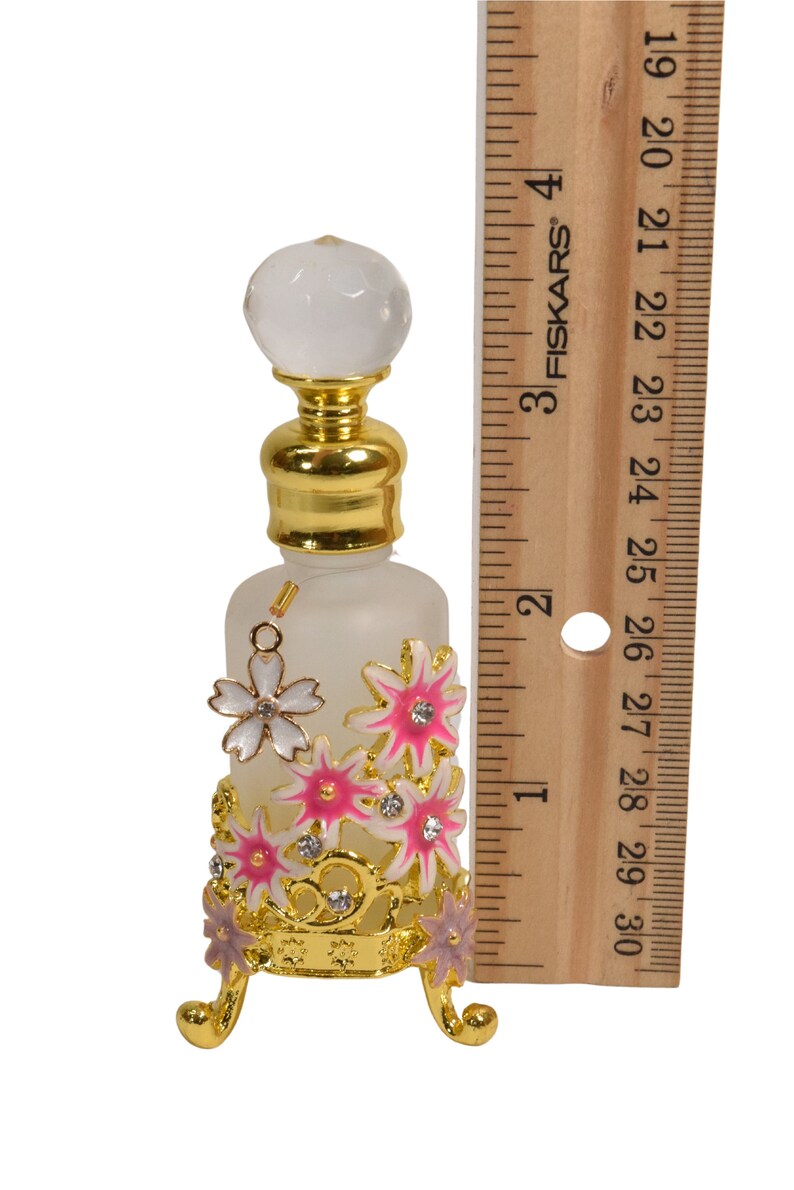 May include: A frosted glass bottle with a gold top and a clear crystal stopper. The bottle is decorated with pink and white flowers and rhinestones. The bottle is sitting on a gold metal stand with three legs.