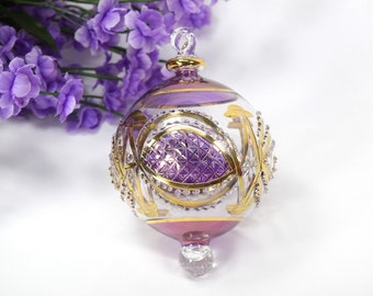 May include: Purple and gold glass Christmas ornament with a geometric design. The ornament is round and has a clear glass top.