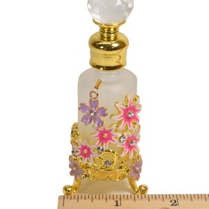 May include: A clear glass bottle with a gold top and a decorative stand. The stand is made of gold metal and features pink and purple flowers with rhinestones. The bottle is approximately 4 inches tall.