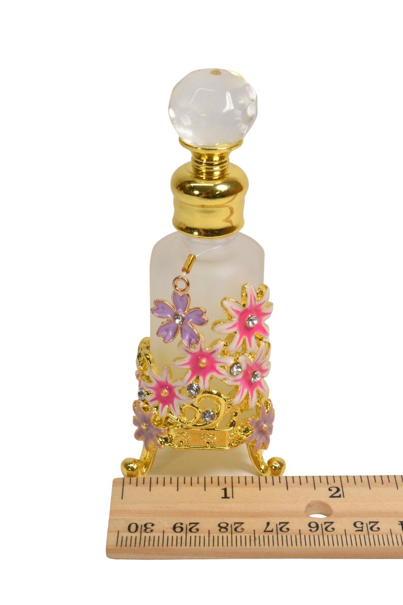 May include: A clear glass bottle with a gold top and a decorative stand. The stand is made of gold metal and features pink and purple flowers with rhinestones. The bottle is approximately 4 inches tall.