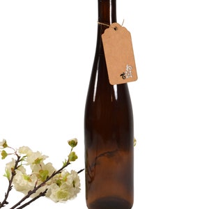 Puede incluir: Una botella de vidrio marr&oacute;n con un tap&oacute;n de corcho y una etiqueta de papel marr&oacute;n con un dise&ntilde;o blanco. La botella est&aacute; sobre un fondo blanco.
