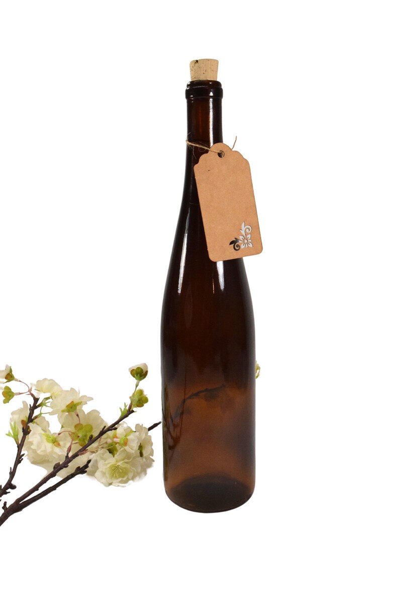 Puede incluir: Una botella de vidrio marr&oacute;n con un tap&oacute;n de corcho y una etiqueta de papel marr&oacute;n con un dise&ntilde;o blanco. La botella est&aacute; sobre un fondo blanco.