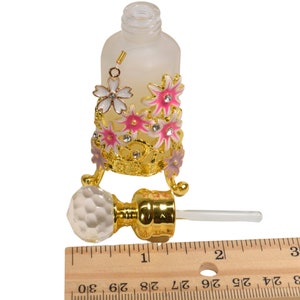 May include: A frosted glass bottle with a gold metal decorative stand featuring pink and white flowers. The bottle has a clear glass dropper with a gold metal top. The bottle is approximately 2 inches tall.