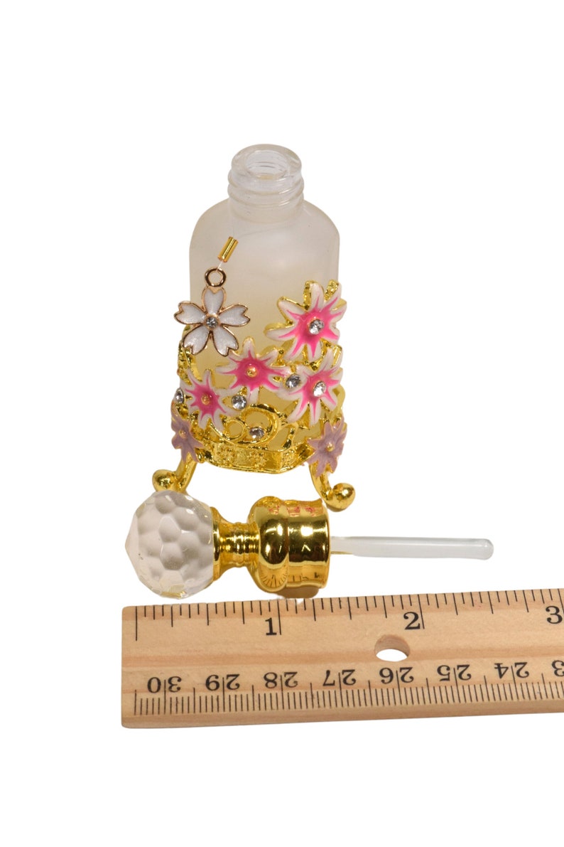 May include: A frosted glass bottle with a gold metal decorative stand featuring pink and white flowers. The bottle has a clear glass dropper with a gold metal top. The bottle is approximately 2 inches tall.