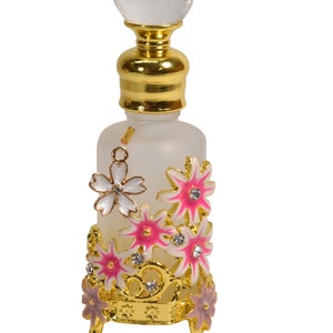 May include: A frosted glass bottle with a gold top and a crystal stopper. The bottle is decorated with pink and white flowers and rhinestones. The bottle sits on a gold stand with three legs.