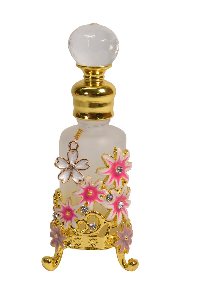 May include: A frosted glass bottle with a gold top and a crystal stopper. The bottle is decorated with pink and white flowers and rhinestones. The bottle sits on a gold stand with three legs.