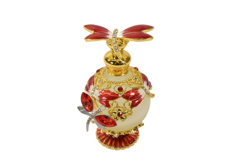 May include: A white glass perfume bottle with a gold and red dragonfly design. The bottle has a round body and a decorative base. The dragonfly is perched on the top of the bottle.