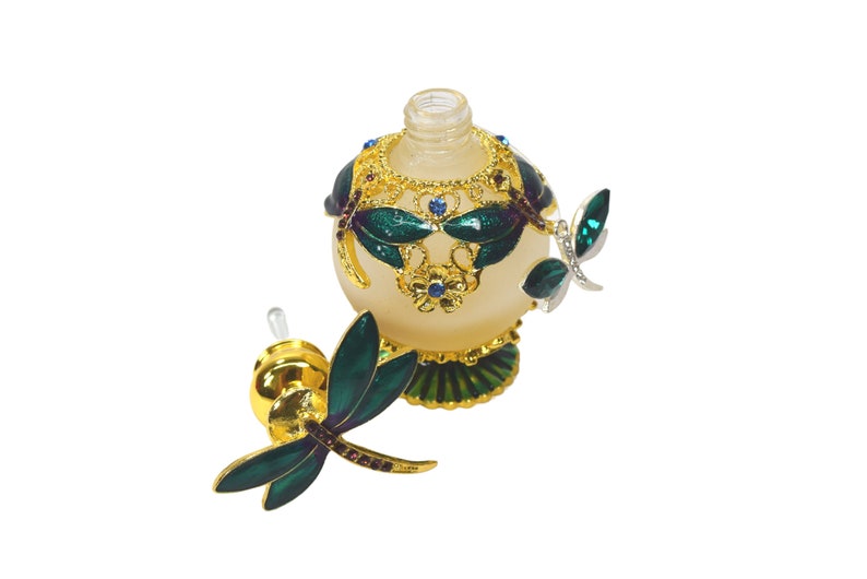 May include: A gold and green glass perfume bottle with a dragonfly design. The bottle has a clear glass stopper with a gold top and a dragonfly design.
