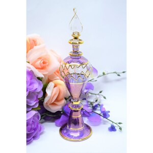 May include: A tall, slender purple glass perfume bottle featuring a clear glass stopper with a twisted finial. The bottle is decorated with horizontal gold bands and a zigzag gold pattern around the bulbous midsection, resting on a circular base.