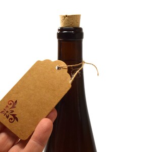 Puede incluir: Una botella marr&oacute;n con un tap&oacute;n de corcho y una etiqueta de regalo de papel marr&oacute;n atada alrededor del cuello con una cuerda. La etiqueta de regalo tiene un dise&ntilde;o floral decorativo.