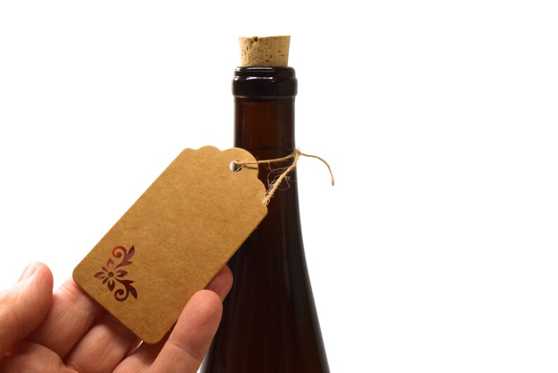 Puede incluir: Una botella marr&oacute;n con un tap&oacute;n de corcho y una etiqueta de regalo de papel marr&oacute;n atada alrededor del cuello con una cuerda. La etiqueta de regalo tiene un dise&ntilde;o floral decorativo.