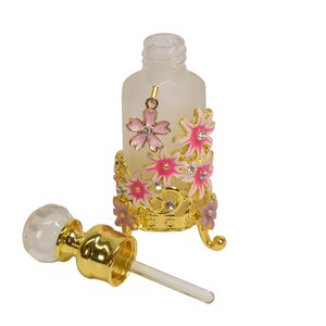 May include: A clear glass bottle with a gold metal floral design and a matching dropper. The bottle is frosted and has a gold cap. The floral design features pink and white flowers with gold accents.
