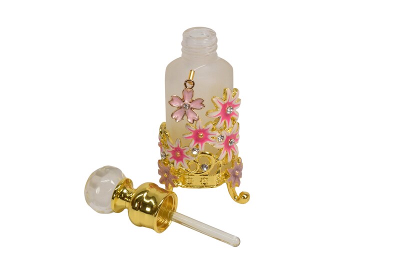 May include: A clear glass bottle with a gold metal floral design and a matching dropper. The bottle is frosted and has a gold cap. The floral design features pink and white flowers with gold accents.