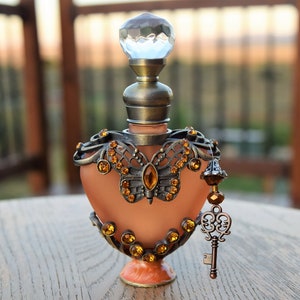 Op de afbeelding: Een sierlijk parfumflesje met een perzikkleurig lichaam en een decoratief vlinderontwerp. De fles heeft een kristallen stop, bronzen accenten en amberkleurige juwelen. Een sleutelbedel hangt aan de zijkant.