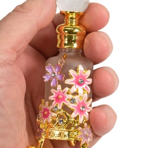 May include: A small glass bottle with a gold top and a clear crystal stopper. The bottle is decorated with pink and purple flowers and gold accents. The bottle is sitting on a gold stand with three legs.
