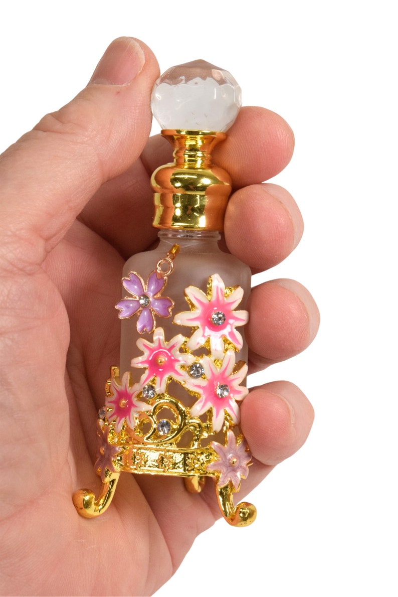 May include: A small glass bottle with a gold top and a clear crystal stopper. The bottle is decorated with pink and purple flowers and gold accents. The bottle is sitting on a gold stand with three legs.