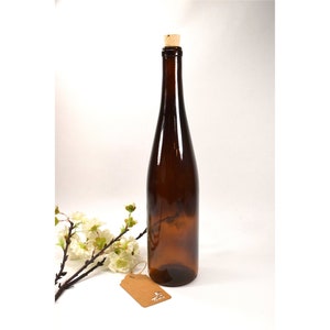 Puede incluir: Una botella de vidrio marr&oacute;n con un tap&oacute;n de corcho. La botella est&aacute; vac&iacute;a y tiene una ligera curva en el cuerpo. La botella est&aacute; sobre una superficie blanca con una peque&ntilde;a etiqueta marr&oacute;n.