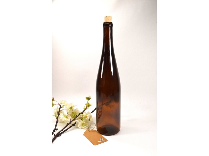 Puede incluir: Una botella de vidrio marr&oacute;n con un tap&oacute;n de corcho. La botella est&aacute; vac&iacute;a y tiene una ligera curva en el cuerpo. La botella est&aacute; sobre una superficie blanca con una peque&ntilde;a etiqueta marr&oacute;n.