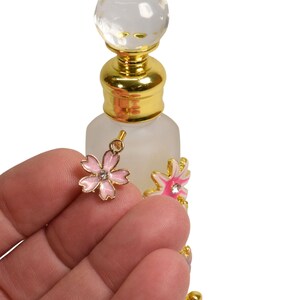 May include: A small, clear glass bottle with a gold top and a crystal ball stopper. The bottle has two pink and white flower charms attached to it.