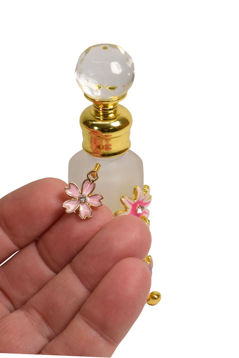May include: A small, clear glass bottle with a gold top and a crystal ball stopper. The bottle has two pink and white flower charms attached to it.