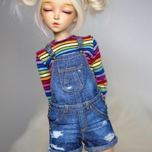 Puede incluir: Una muñeca articulada de piel pálida y cabello rubio recogido en dos moños, con una diadema blanca de orejas de gato. La muñeca viste una camiseta de manga larga con rayas horizontales de arcoíris bajo un peto corto de mezclilla azul desgastado.