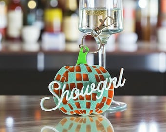 May include: A decorative keychain with a pumpkin-shaped design. The pumpkin has a mosaic pattern in teal and orange, with the word "Showgirl" in white script. The keychain is attached to a silver clasp and ring, with a green stem.