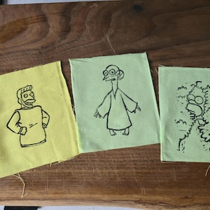 Könnte beinhalten: Drei Stoffflicken mit schwarzen Strichzeichnungen von Zeichentrickfiguren. Der linke Flicken ist gelb, der mittlere hellgrün und der rechte hellgrün. Die Charaktere stammen aus einer beliebten Zeichentrickserie.