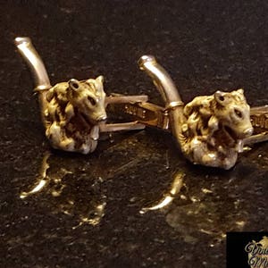 May include: A pair of gold cufflinks featuring a detailed sculpted design of a boar's head. The cufflinks are made of metal and have a polished finish.