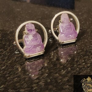 May include: A pair of silver cufflinks with amethyst stone carvings of a seated Buddha. The cufflinks are circular and have a polished silver finish.