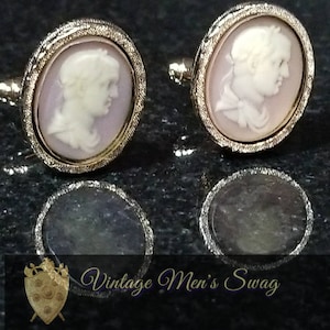 Op de afbeelding: Een paar gouden manchetknopen met een cameo-ontwerp. Elke manchetknoop heeft een wit cameo-portret van een persoon op een lichtpaarse achtergrond.