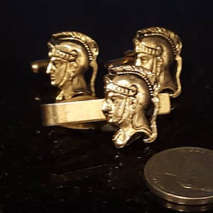 May include: Three gold-toned cufflinks featuring a Roman soldier design. Each cufflink has a detailed depiction of a soldier's head wearing a helmet.