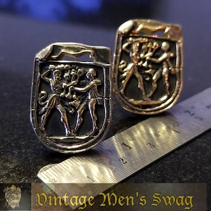 May include: Two gold-toned cufflinks with a shield-shaped design. Each cufflink features a raised image of two figures holding a staff with a crown on top. The cufflinks are on a ruler with markings in inches.