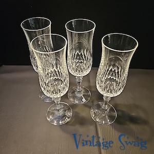 May include: Four clear crystal champagne flutes. Each flute has a diamond-cut pattern on the bowl and a long stem with a circular base. The flutes are arranged on a dark surface.