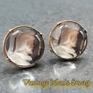May include: A pair of vintage cufflinks with a cameo design. The cufflinks are gold-toned and feature a clear glass dome over a white cameo image of a man's profile.