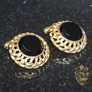 Könnte beinhalten: Ein Paar goldfarbene Manschettenknöpfe mit einem schwarzen, oval geformten Stein in der Mitte. Die goldene Fassung hat ein offenes, filigranes Design. Die Manschettenknöpfe werden auf einer dunklen, strukturierten Oberfläche präsentiert. Der Text "Vintage Men's Swag" ist enthalten.