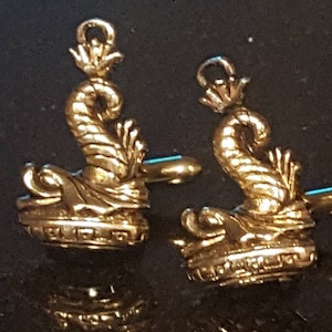 Könnte beinhalten: Ein Paar goldfarbene Manschettenknöpfe mit einem detaillierten Design eines stilisierten Seepferds mit einer Krone auf dem Kopf.