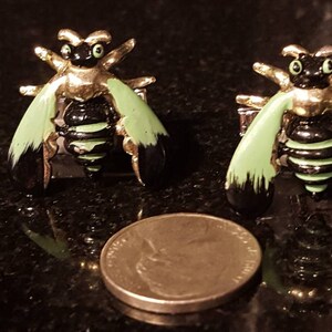 Può includere: Un paio di gemelli a forma di ape. I gemelli presentano un corpo color oro, un addome a strisce nere e verdi e ali verdi e nere. Una moneta è accanto ai gemelli per il confronto delle dimensioni.