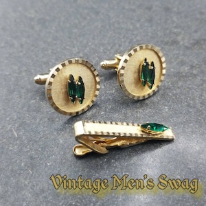 May include: A set of gold cufflinks and a tie clip with green gemstone accents. The cufflinks are round and have a textured surface. The tie clip is rectangular and has a textured surface. The gemstones are emerald green and have a marquise shape.