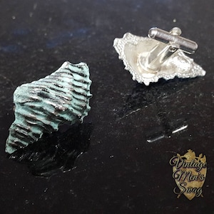 May include: A pair of cufflinks, one is a silver seashell design, the other is a textured green seashell.