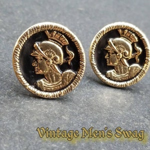 May include: A pair of gold-toned cufflinks with a black background and a raised image of a person wearing a helmet. The cufflinks have a textured edge.