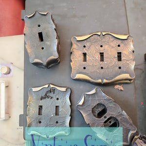 May include: A collection of four dark bronze-toned metal light switch and outlet wall plates with a textured, cracked surface finish. The set includes one single toggle plate, one triple toggle plate, one double toggle plate, and one duplex outlet plate.