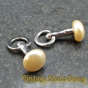 May include: A pair of silver cufflinks with a pearl on each end. The cufflinks have a circular loop on one end and a small post on the other end.