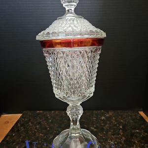 May include: A clear, vintage glass candy dish with a diamond pattern and a red band. The dish has a lid with a pointed finial and sits on a pedestal base. The words "Vintage Swag" are at the bottom.